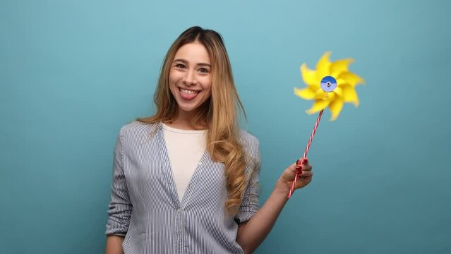 Portrait of carefree wavy haired woman blowing at paper windmill, playing with pinwheel toy on stick, happy expression, wearing striped shirt. Indoor studio shot isolated on blue background.