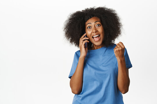 Enthusiastic Black Woman Answer Phone Call And Celebrating Victory, Hear Great News During Mobile Conversation, Standing In Blue Tshirt Over White Background