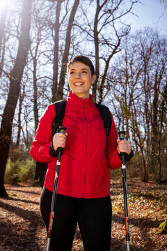 The smiling young woman is hiking in the woods. The woman has short hair, wears a red jacket and uses trekking poles. Travel and outdoors concept.