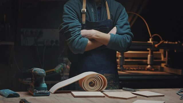 Portrait of an unidentifiable male craftsman in his workshop. A man in a working apron stands with his arms folded across his chest near the desktop on which carpentry tools and wooden blanks lie