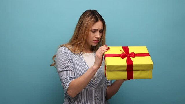 Portrait of upset displeased young adult woman holding gift box, opening present with dissatisfied expression, wearing striped shirt. Indoor studio shot isolated on blue background.