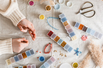The girl's hands are weaving a beaded bracelet and items for beading on the table. Development of creative skills and fine motor skills for children. Top view. - obrazy, fototapety, plakaty