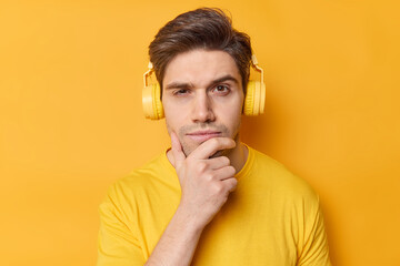 Obraz na płótnie Canvas Horizontal shot of thoughtful man holds chin and looks seriously at camera listens favorite song via stereo headphones dressed in casual clothes poses against vivid yellow background. Let me think