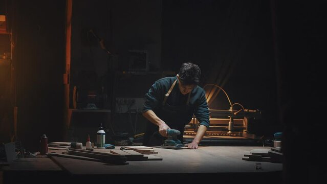 General plan cinematic video of a carpenter at work in a carpentry workshop. The guy polishes wooden parts with a grinder. Dark key, male hobby, family carpentry business