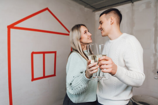 Couple In Unfinished Building Celebrating Moving To A New Flat. Couple Drinking Champagne - Cheers. Young Couple In New Property Dreaming About Renovation Near Red Drawn House On A Wall