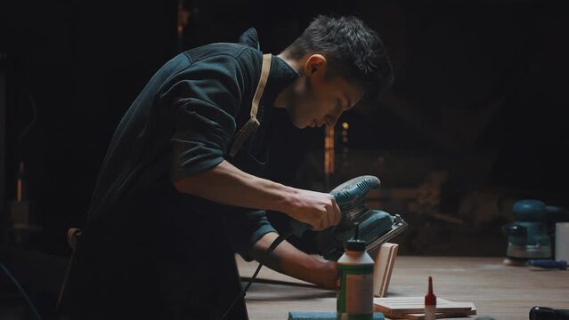 Side view cinematic video of a carpenter at work in a carpentry workshop. The guy polishes wooden parts with a grinder. Dark key, male hobby, family carpentry business