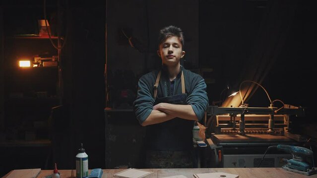 Confident young craftsman stands near a work table in a carpentry workshop. A man in a working apron shakes his hands from chips and crosses his arms over his chest, general plan