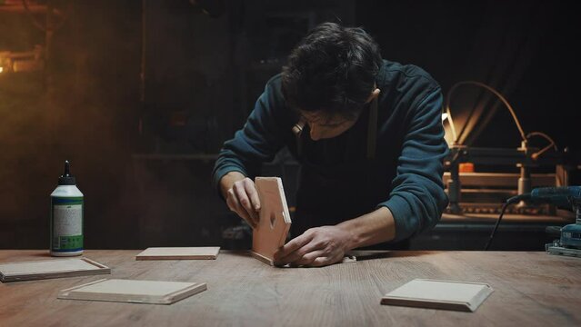 Attractive brunette carpenter is gluing together wooden blanks for a birdhouse. Male hobby, family business concept. Cinematic effect