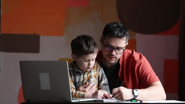 Father is working in home office with a son. At table with laptop computer