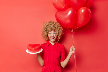Horizontal shot of surprised positive young woman gets present from boyfriend on Valentines Day holds heart shaped cake and balloons wears dress enjoys favorite holiday isolated over red studio wall