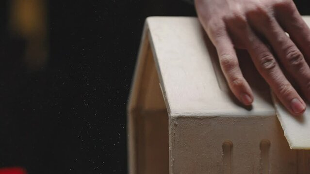 Carpenter polishing the corners of a wooden bird house, carpentry workshop, close-up of the craftsman's hand. Cinematic effect, dark key, sawdust and dust in the air are visible, slow motion