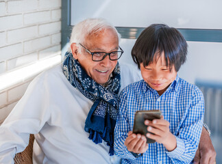 a white-haired caucasian grandfather and his dark-haired asian grandson play with the mobile phone on the terrace. they are laughing and showing happiness. concept adoption and family.