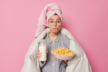 Young woman going to have breakfast holds bowl of cereals and millk bottle wrapped in duvet wears pajama spectacles and bath towel on head awakes early in morning isolated on pink background