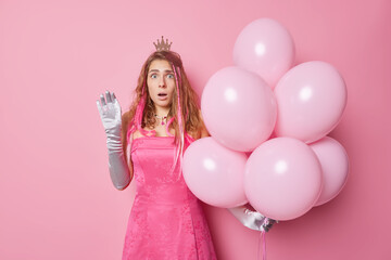 Obraz na płótnie Canvas Shocked stunned young woman wears festive dress and gloves holds big bunch of helium balloons comes on birthday party reacts on something amazing isolated over pink background. Holiday concept