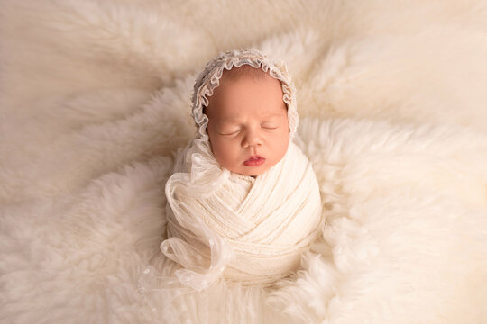 Sleeping newborn girl in the first days of life in a white cocoon with a white cap on a white background. Studio macro photography, portrait of a newborn. The concept of female happiness.