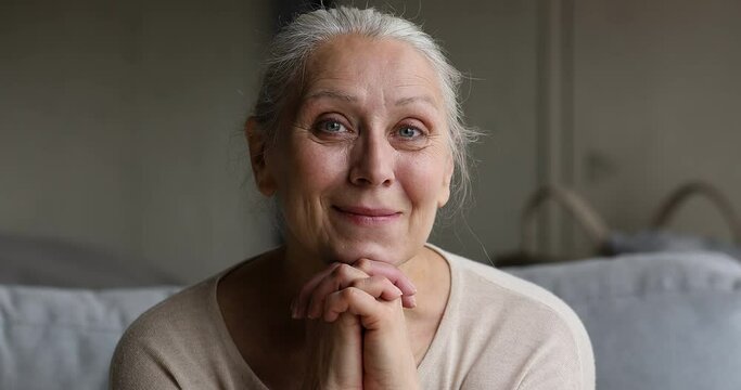 Close up kind older good-looking grey-haired woman sits on sofa looks into distance smiling while remembering past with warmth and gratitude. Carefree life of retiree on retirement, well-being concept