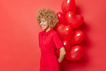 Horizontal shot of cheerful curly haired woman prepares surprise for lover on Valentines Day holds bunch of heart balloons behind back smiles happily wears dress isolated over vivid red background