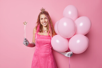 Obraz na płótnie Canvas Horizontal shot of cheerful woman prepares for celebration wears festive dress long gloves crown holds magic wand and bunch of inflated balloons isolated over pink background. Holidays concept