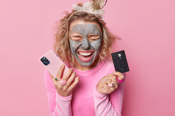 Emotional young woman screams loudly keeps eyes closed reacts on something holds mobile phone and credit card makes shopping online applies beauty mask on face isolated over pink background.