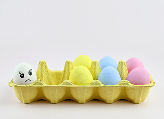 A white chicken egg with a sad emotion drawn stands in front of multi-colored eggs. The concept of...