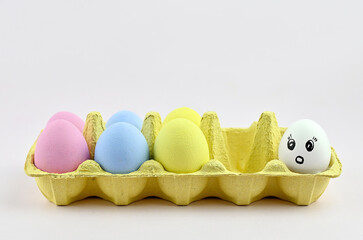 A tray with colored Easter eggs and a white egg next to it with a surprised facial expression. The concept of social status. The concept is not like everyone else, different from others.