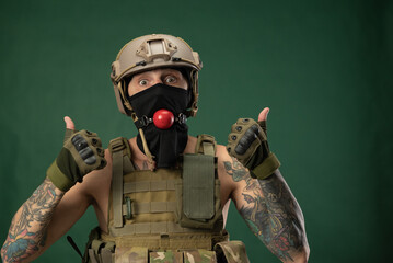 a soldier man in military clothes helmet with a bdsm gag in his mouth expresses emotions, photo joke - obrazy, fototapety, plakaty