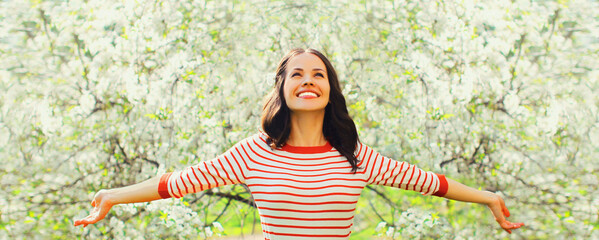 Happy smiling young woman raising her hands up in spring blooming garden on flowers background