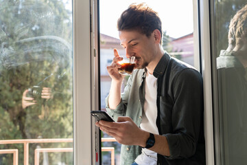 One young caucasian man having cup of coffee while standing by the window at home in sunny day wearing shirt browsing internet on mobile phone sending text messages in morning real people copy space - Powered by Adobe