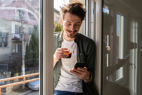 One young caucasian man having cup of coffee while standing by the window at home in sunny day wearing shirt browsing internet on mobile phone sending text messages in morning real people copy space