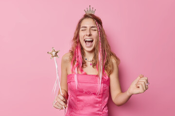 Happy woman with long hair exclaims loudly clenches fist keeps mouth widely opened holds magic wand wears dress and crown celebrates something isolated over pink background. Holiday concept.