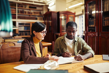Portrait of mature female professor tutoring African-American student in classic library interior...