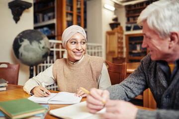 Portrait of smiling Muslim woman wearing head covering while studying in college library with...