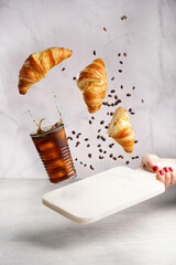 A glass with black iced coffee, coffee beans and freshly baked croissants flying in the air on white background. Food levitation