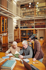 Vertical portrait of white haired senior man studying in library with diverse group of students,...