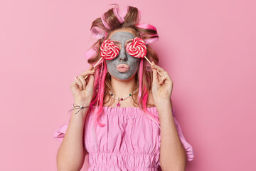 Photo of young woman applies beauty mask covers eyes with caramel candies makes hairstyle dressed in blouse isolated over pink background. Feminity hairstyling skin care and sweet tooth concept
