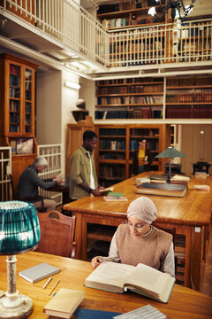Vertical wide angle shot of classic college library with diverse group of people, focus on young woman wearing head covering and reading book