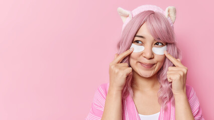 Horizontal shot of Asian woman with pink dyed hair points at beauty patches under eyes to remove wrinkles looks thoughtfully away wears soft headband isolated over pink background blank space
