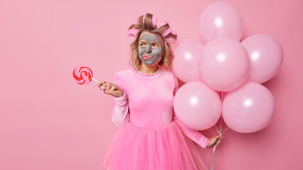 Displeased angry young woman looks thoughtfully away applies beauty mask on face undergoes beauty procedures before special occasion holds caramel candy and inflated balloons pink background
