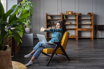 Happy satisfied with work done woman stretching hands, relaxing in cozy armchair with laptop in...