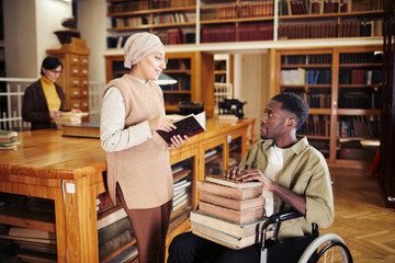 Portrait of young African-American man in wheelchair holding stack of books and chatting with...