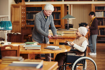 Portrait of smiling senior professor helping young woman in wheelchair at college library