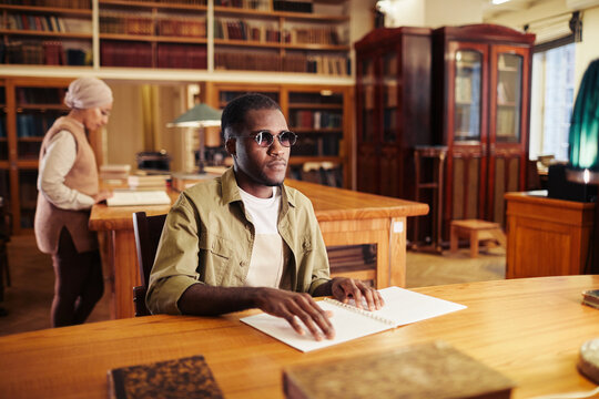 Portrait of young black man with vision impairment reading book in braille at table in college library