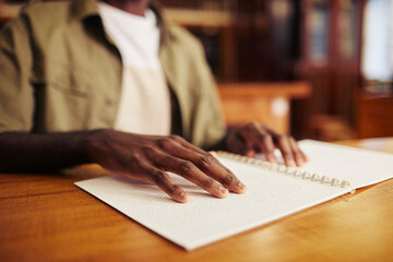 Close up of young black man with vision impairment reading book in braille at table in college...