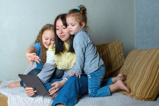 The Older Sister Is Talking And Watching Something Interesting On The Tablet, The Younger Two Sisters Are Interested In Watching What Is On The Screen Of The Gadget. Family Fun At Home