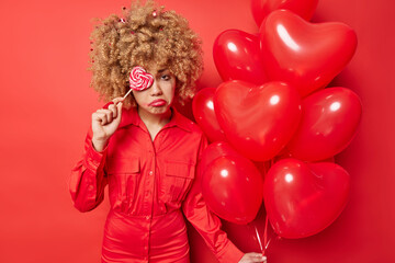 Discontent curly haired woman feels frustrated and lonely on Valentines Dy holds heart balloons covers eye with delicious candy wears dress isolated over vivid red background. Bad mood celebration