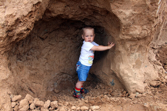 A little boy with blond hair on a walk outside on a summer day. The boy is sitting in a clay cave.
