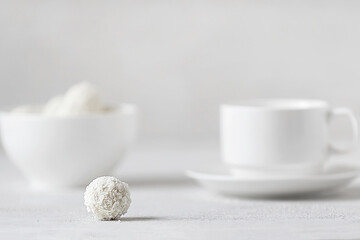 on a white background lies one round candy in coconut flakes on the background in the blur zone a vase of sweets and a mug of tea. selective focus