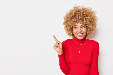 Positive curly haired woman points finger on left side shows blank space for your advertising content wears casual red turtleneck isolated over white background. Place your company title here