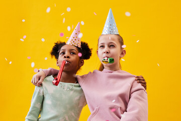 Portrait of two girls wearing party hats while celebrating Birthday against pop yellow background...