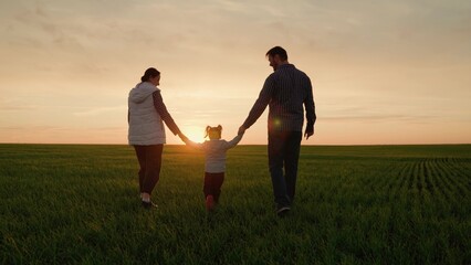 Happy family, dad daughter mom walk together on green grass at sunset. Father mother kid girl walk in park holding hands in sun. Child parents are walking along green field. Family hold hands teamwork